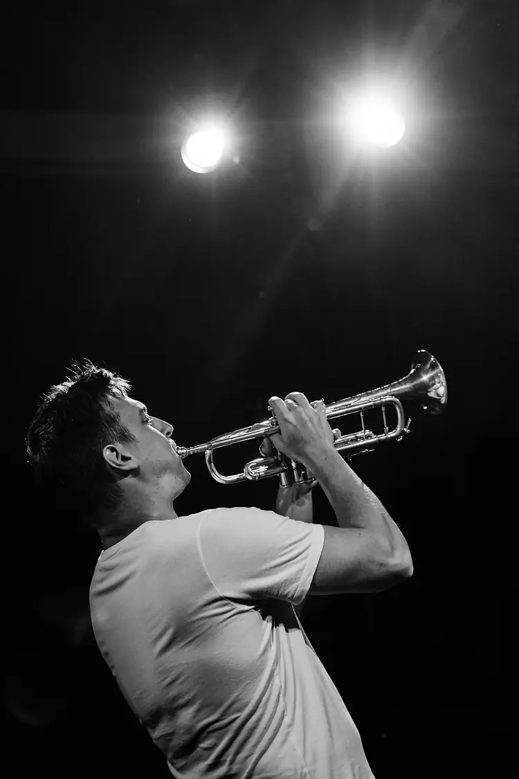 Trumpeter lifting his instrument toward two bright stage lights, in black and white — Jazz photography by Michaël Gramm